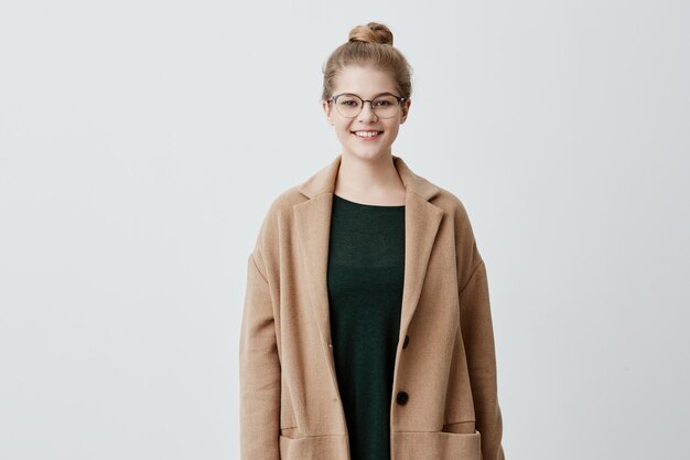 Heureuse jolie femme aux cheveux blonds en noeud, lunettes et peau saine vêtue d'un manteau brun sur un pull vert souriant tout en posant contre le mur de béton. Personnes et style de vie