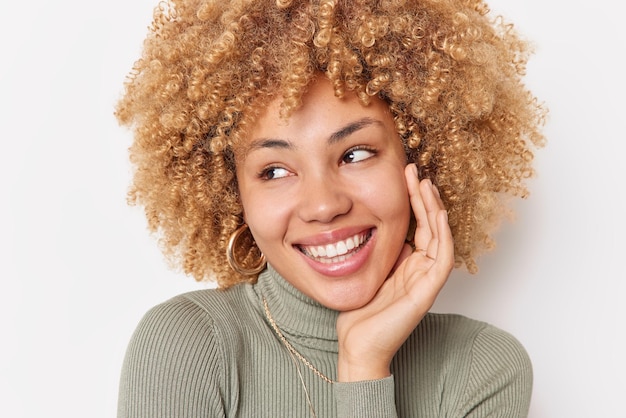 Photo gratuite heureuse jolie femme aux cheveux bouclés garde la main sur la joue regarde au loin les sourires porte joyeusement un col roulé décontracté étant de bonne humeur isolé sur fond blanc concept de personnes et d'émotions positives