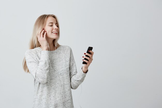 Heureuse jolie femme aux cheveux teints blonds debout à l'intérieur avec son smartphone chat