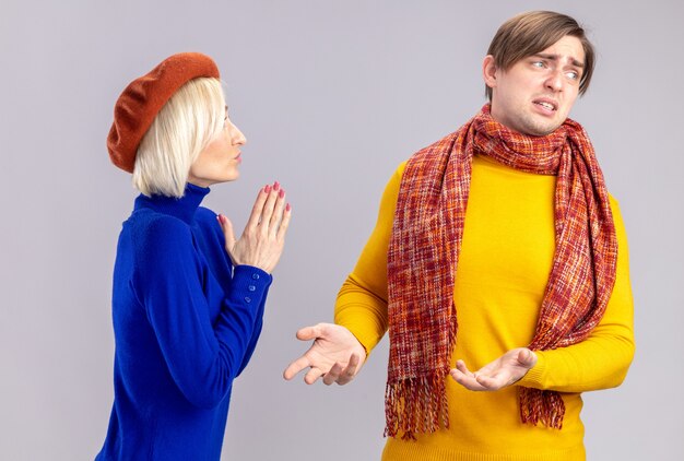 Heureuse jolie femme blonde avec béret tenant les mains ensemble et regardant un bel homme slave mécontent avec un foulard autour du cou isolé sur un mur blanc avec espace de copie