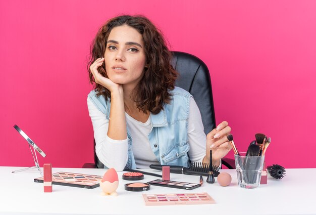 Heureuse jolie femme caucasienne assise à table avec des outils de maquillage mettant la main sur le menton et isolée sur un mur rose avec espace de copie