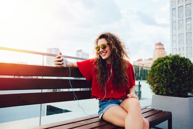 Heureuse jolie fille aux cheveux bouclés, dans les écouteurs, regarde l&#39;écran du smartphone