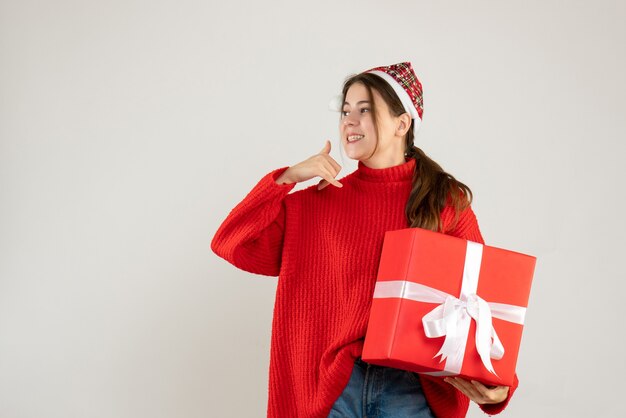 Heureuse jolie fille avec bonnet de noel tenant un cadeau lourd faisant appelez-moi signe debout sur blanc