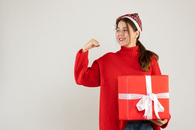 heureuse jolie fille avec bonnet de noel tenant un cadeau lourd montrant sa force debout sur blanc