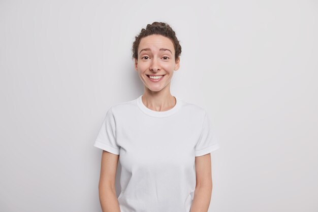 Heureuse jolie fille millénaire avec des sourires de beauté naturelle a joyeusement les cheveux peignés foncés vêtus de modèles de t-shirt décontractés à l'intérieur sur blanc exprime des sentiments sincères écoute smth intéressant