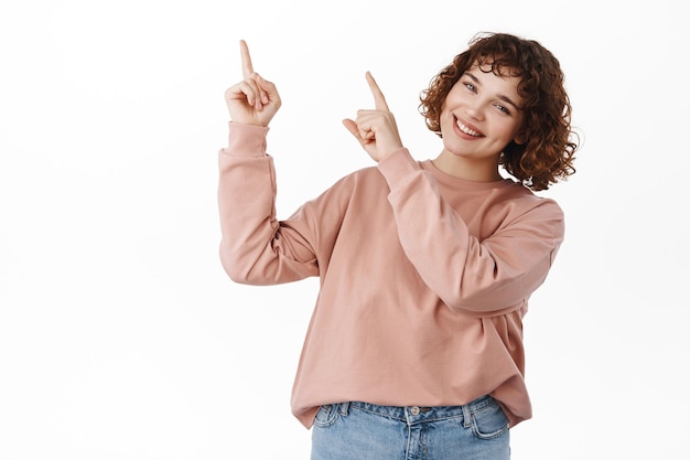 Heureuse jolie fille pointant vers la bannière du coin supérieur gauche, montrant le logo avec des informations d'annonce ou de réduction, souriant largement à la caméra, debout sur fond blanc.