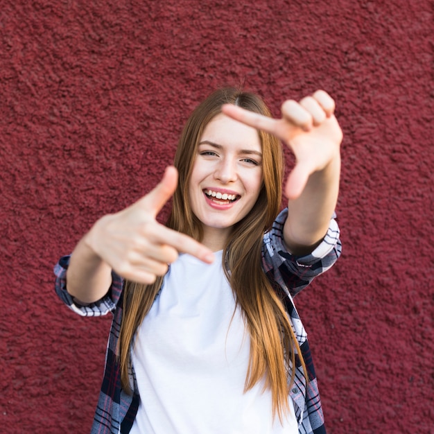 Photo gratuite heureuse jolie jeune femme faisant le cadre de la main contre le mur rouge