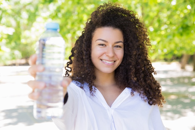 Heureuse jolie jeune femme montrant une bouteille d'eau dans le parc