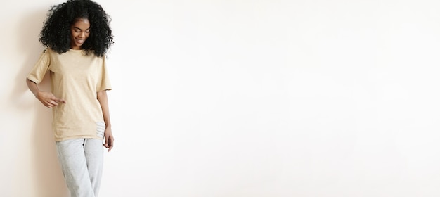 Heureuse Jolie Jeune Fille Africaine Avec Une Coiffure Afro Vêtue D'un T-shirt Surdimensionné Vierge, Pointant Le Doigt Dessus Et Souriant Joyeusement