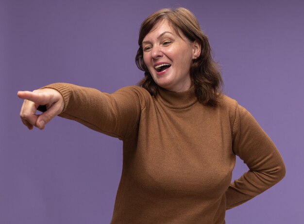 Heureuse et joyeuse femme d'âge moyen en col roulé marron à côté en pointant avec l'index à quelque chose debout sur le mur violet