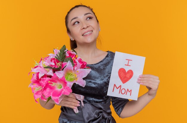 Heureuse et joyeuse femme asiatique mère tenant une carte de voeux et un bouquet de fleurs célébrant la fête des mères à la recherche de sourire joyeusement debout sur le mur orange