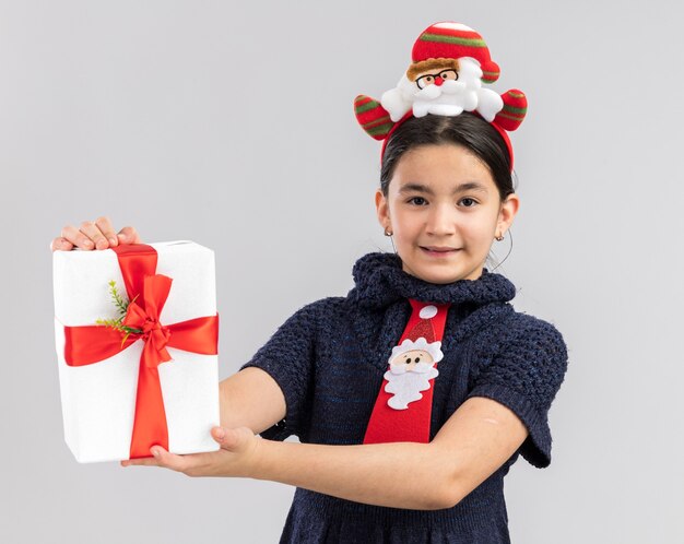 Heureuse et joyeuse petite fille en robe en tricot portant une cravate rouge avec une jante de Noël drôle sur la tête tenant un cadeau de Noël à la recherche avec le sourire sur le visage