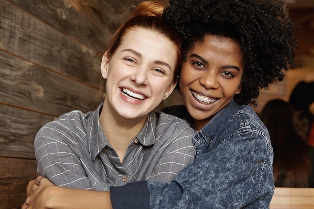 Heureuse lesbienne afro-américaine élégante avec des accolades et des cheveux bouclés tenant fermement sa belle petite amie rousse