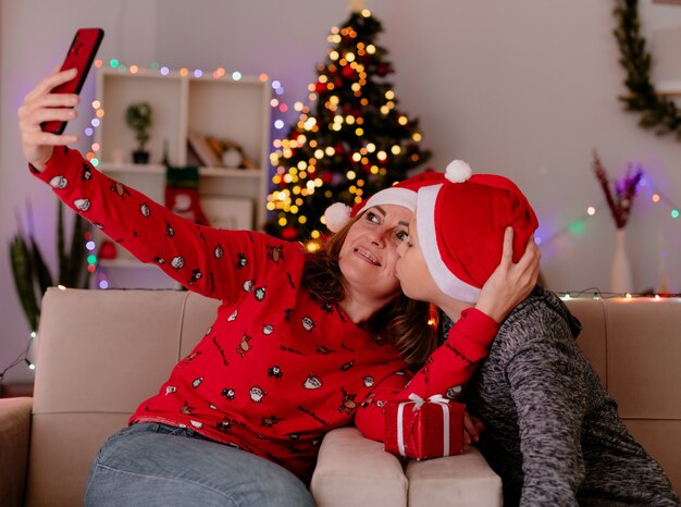 Heureuse mère en bonnet de Noel et petit fils en bonnet de noel avec un cadeau embrassant la mère assise sur un canapé faisant selfie à l'aide de smartphone s'amusant dans une salle décorée avec arbre de Noël en arrière-plan