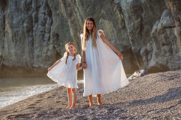 Heureuse mère et fille en robe blanche debout et tenant leurs robes en bord de mer pendant le coucher du soleil, vue de face.