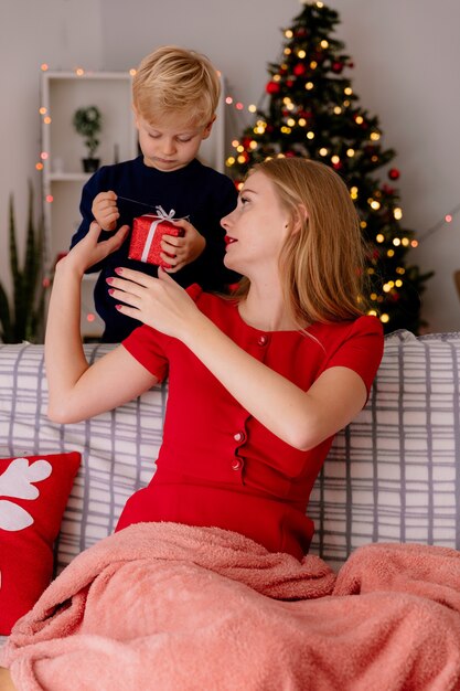 Heureuse mère en robe rouge assise sur un canapé souriant tandis que son petit enfant debout derrière donne un cadeau à sa mère dans une pièce décorée avec un arbre de Noël dans le mur