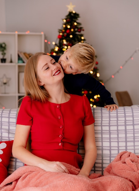 Heureuse mère en robe rouge assise sur un canapé souriant tandis que son petit enfant debout derrière embrassant sa mère dans une pièce décorée avec un arbre de Noël dans le mur