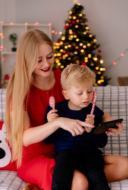 Heureuse mère en robe rouge avec son petit enfant assis sur un canapé avec smartphone montrant quelque chose dans une pièce décorée avec arbre de Noël en arrière-plan