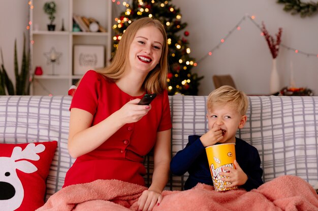 Heureuse mère en robe rouge avec son petit enfant assis sur un canapé sous une couverture avec un seau de pop-corn regardant la télévision ensemble dans une pièce décorée avec un arbre de Noël dans le mur