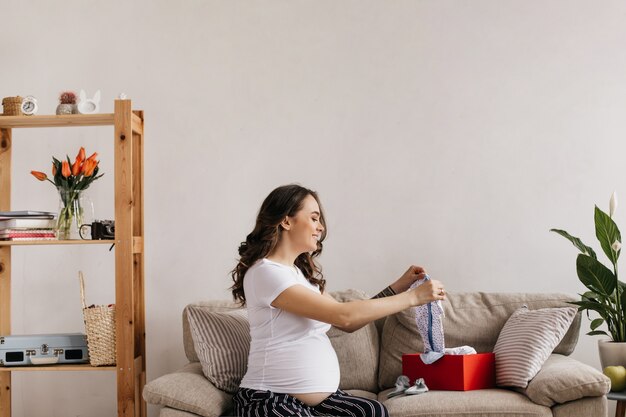 Heureuse mère en tee-shirt blanc et pantalon se penche sur ses futurs vêtements de bébé