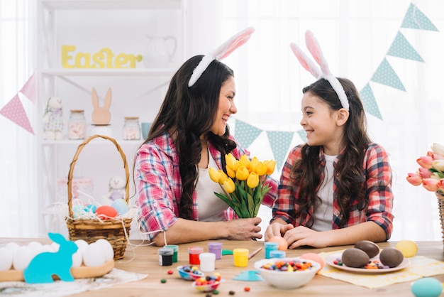 Heureuse mère tenant le bouquet de tulipes en regardant sa fille avec des oeufs de Pâques sur table