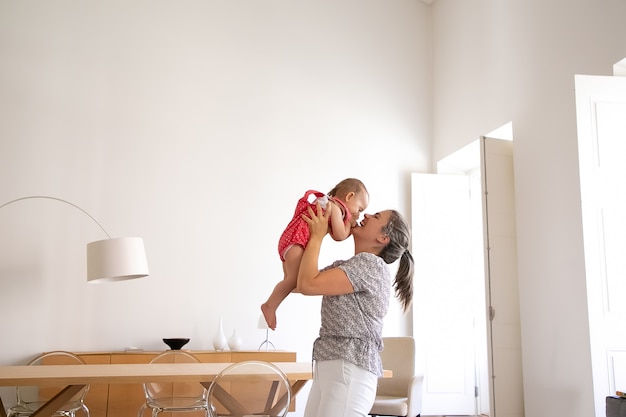 Photo gratuite heureuse mère tenant petite fille, la levant et riant. funny baby girl s'amuser avec maman aimante à l'intérieur et fermer le visage avec des paumes. temps en famille, maternité et concept d'être à la maison