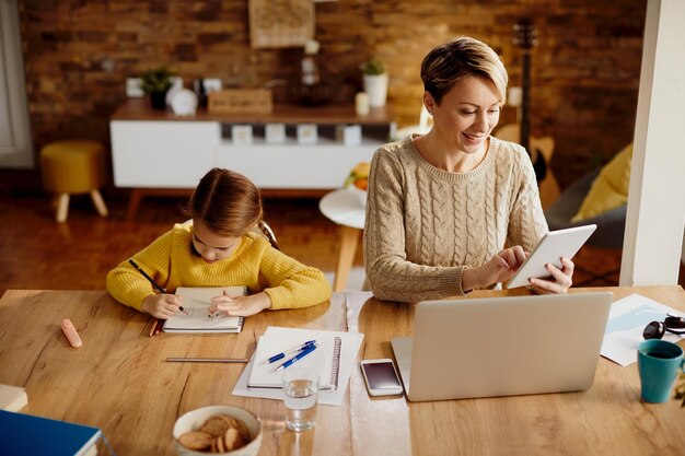 Heureuse mère utilisant un pavé tactile et un ordinateur portable tout en étant avec sa fille à la maison