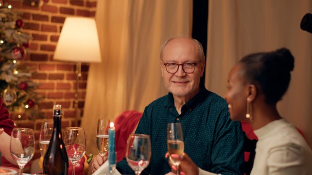 Heureuse personne âgée souriant chaleureusement à la caméra tout en profitant du dîner de Noël avec des parents proches à la maison. Homme âgé joyeux dans la salle à manger célébrant la fête d'hiver traditionnelle. Prise de vue à main levée