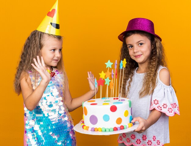 heureuse petite fille blonde avec une casquette de fête debout avec les mains levées et regardant une petite fille caucasienne avec un chapeau de fête violet tenant un gâteau d'anniversaire isolé sur un mur orange avec un espace de copie