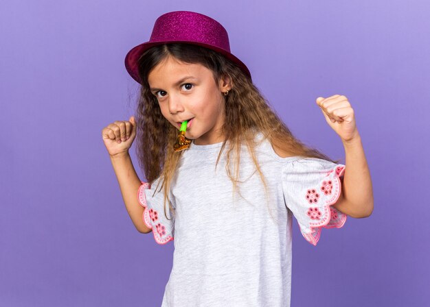 heureuse petite fille caucasienne avec un chapeau de fête violet soufflant un sifflet de fête et gardant les poings isolés sur un mur violet avec espace de copie