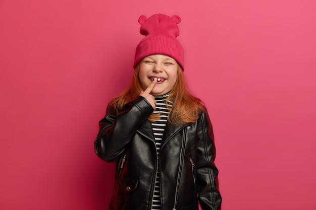 Heureuse petite fille rousse en vêtements à la mode, indique à sa nouvelle dent, a une enfance inoubliable, plisse le visage avec plaisir, pose contre un mur rose vif. Concept de croissance des enfants