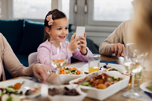 Heureuse petite fille utilisant un téléphone intelligent tout en déjeunant avec sa famille à la maison