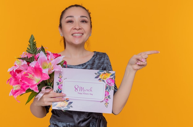 Heureuse et positive femme asiatique mère tenant une carte de voeux et un bouquet de fleurs célébrant la journée internationale de la femme mars