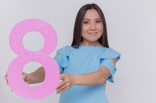 Heureuse et positive femme asiatique tenant le numéro huit en carton à l'avant souriant joyeusement célébrant la journée internationale de la femme debout sur un mur blanc