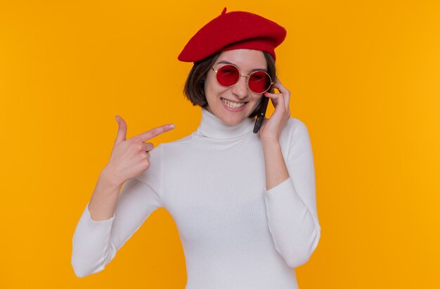 Heureuse et positive jeune femme aux cheveux courts en col roulé blanc portant un béret et des lunettes de soleil rouges parlant au téléphone mobile pointant avec l'index au téléphone mobile debout sur un mur orange