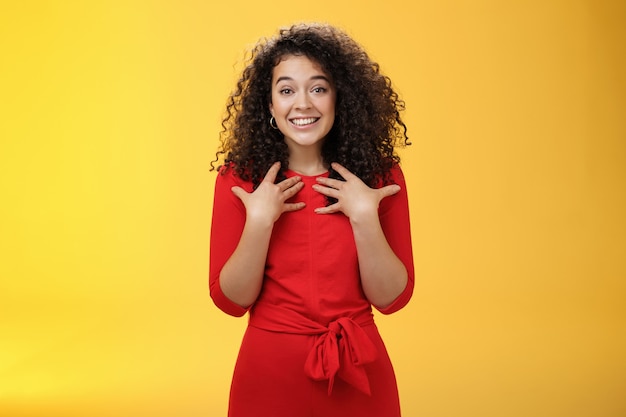 Heureuse et reconnaissante tendre fille gentille aux cheveux bouclés en robe rouge pressant les mains sur la poitrine étant surprise par un cadeau réconfortant tendre et inattendu remerciant le sourire ravi sur fond jaune