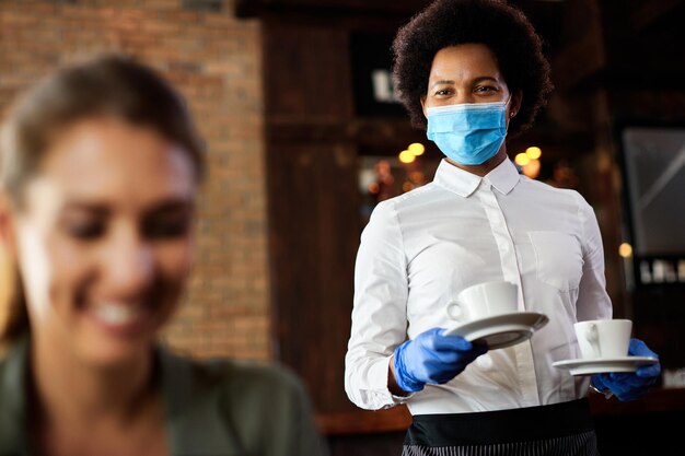 Heureuse serveuse noire avec masque protecteur et gants servant du café dans un café