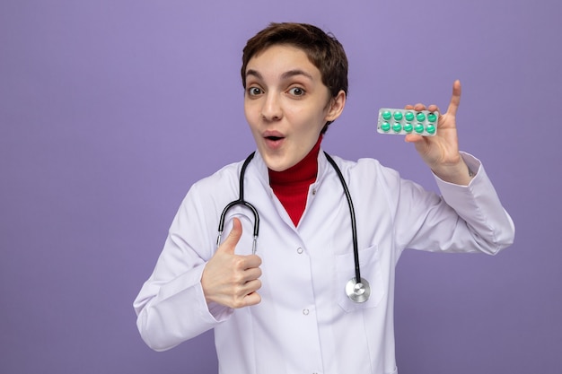 Photo gratuite heureuse et surprise jeune femme médecin en blouse blanche avec stéthoscope tenant un blister avec des pilules à sourire joyeusement montrant les pouces vers le haut