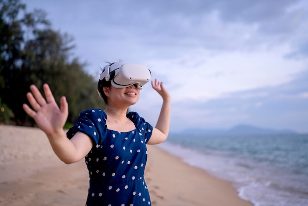 Heureuse tendre gaie attrayante femme adulte asiatique tissu décontracté portant des lunettes de technologie VR debout sur la plage et dispositif de réglage pour visualiser la vidéo numérique virtuelle de simulation 3D sur la vraie nature