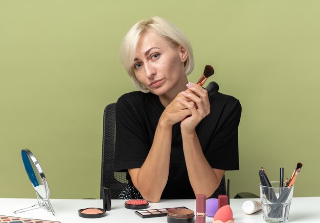 Heureuse tête inclinable jeune belle fille assise à table avec des outils de maquillage tenant des pinceaux à poudre isolés sur un mur vert olive