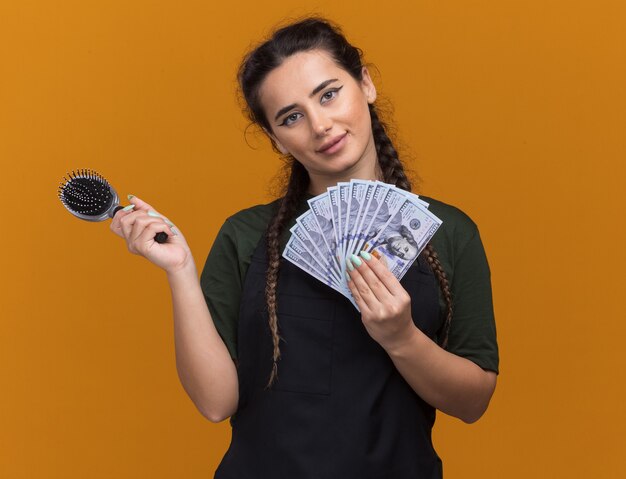 Heureuse tête inclinable jeune coiffeuse en uniforme tenant un peigne avec de l'argent isolé sur un mur orange