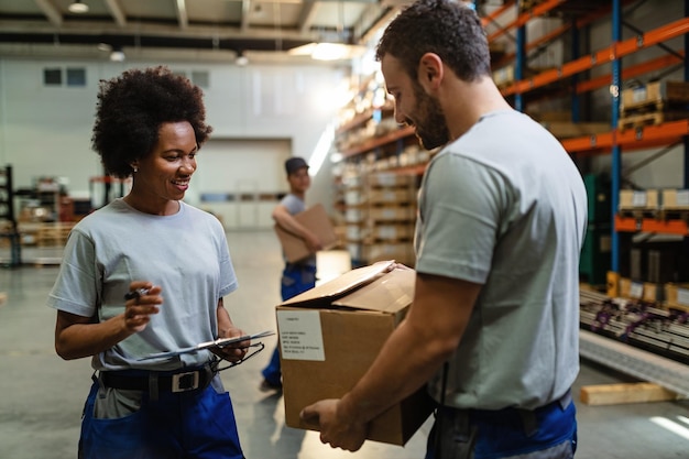 Heureuse travailleuse noire passant par la liste de contrôle et parlant à son collègue qui porte une boîte en carton dans l'entrepôt de distribution