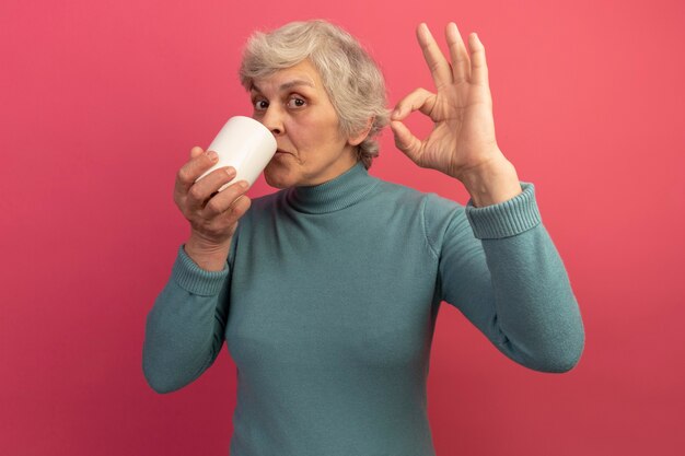 Heureuse vieille femme portant un pull à col roulé bleu buvant une tasse de thé faisant signe ok isolé sur mur rose