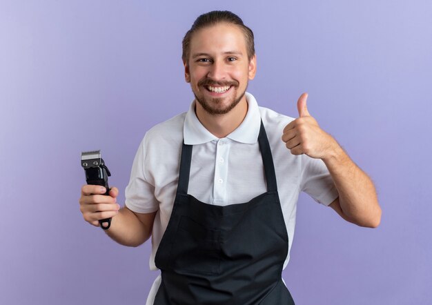 Heureusement souriant jeune beau coiffeur en uniforme tenant une tondeuse à cheveux et montrant le pouce vers le haut isolé sur violet