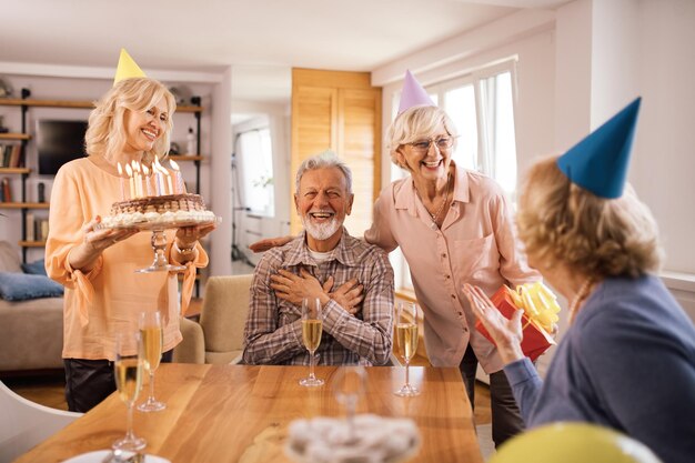 Heureuses femmes âgées surprenant leur ami avec un gâteau pour son anniversaire à la maison