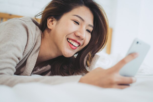 Heureuses femmes asiatiques utilisent un téléphone intelligent sur le lit le matin