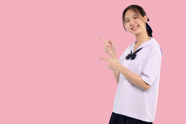 Heureux accolades étudiante asiatique fille en uniforme scolaire doigt pointant sur fond pastel isolé