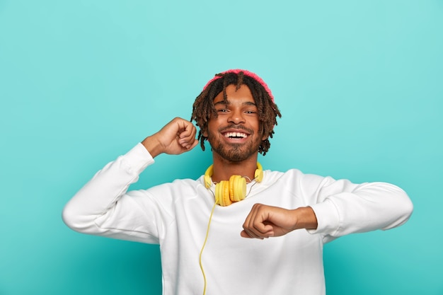 Photo gratuite heureux adolescent a de courtes dreadlocks, se déplace au rythme de la musique, ressent du plaisir, habillé en pull blanc, isolé sur fond bleu