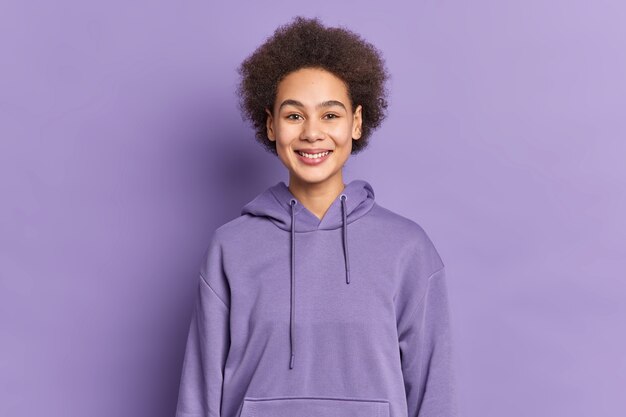 Heureux adolescent ethnique avec des sourires de cheveux afro porte positivement un sweat à capuche violet étant de bonne humeur.