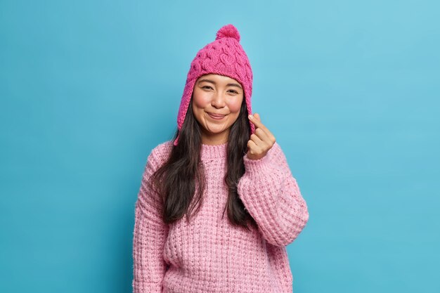 Heureux adorable femme asiatique romantique rend coréen comme des formes de signe mini coeur à l'avant porte un chapeau tricoté et un pull pose contre le mur bleu du studio